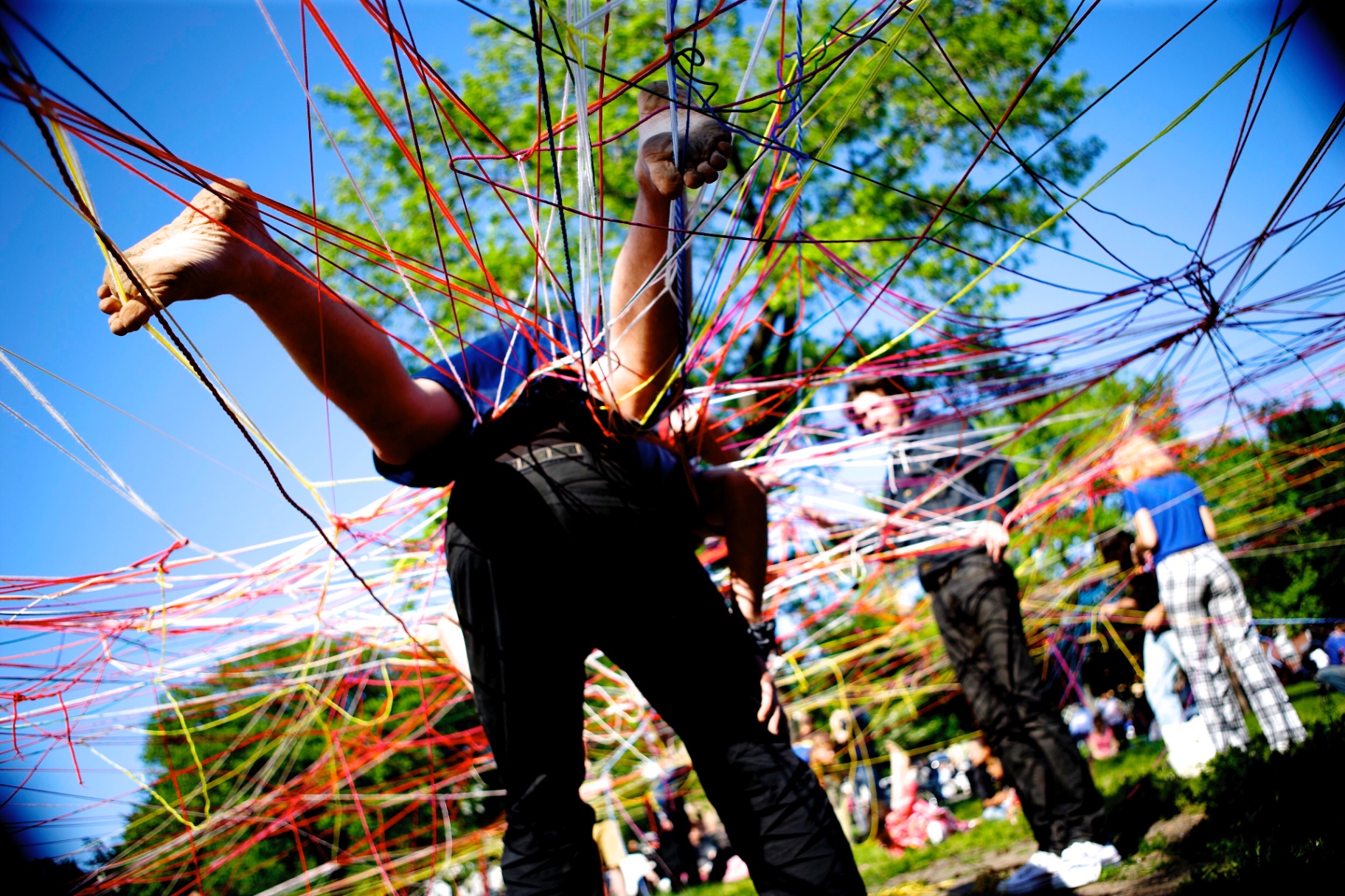 A visitor suspended horizontally inside a rainbow string web while others stand on the lawn below