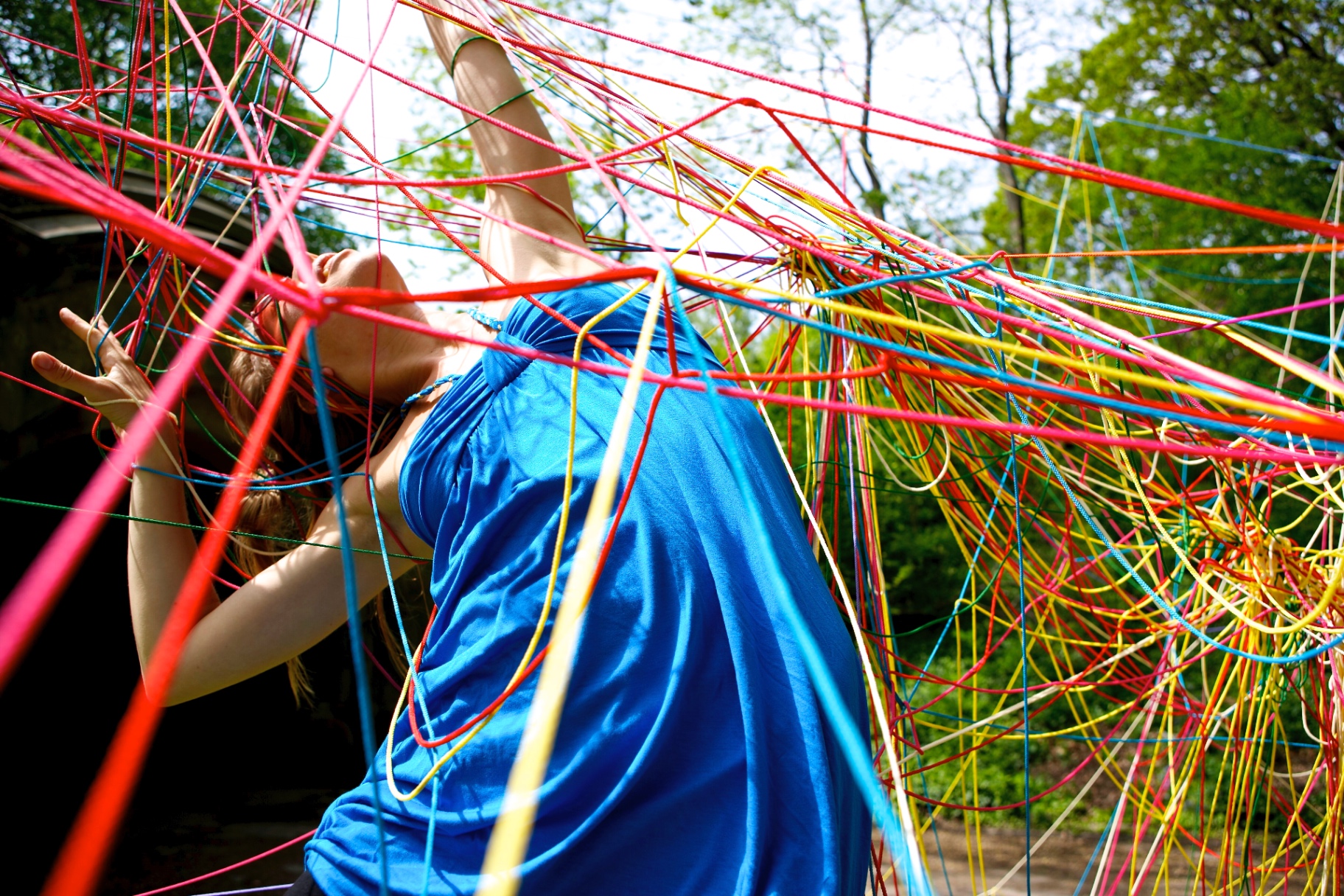 A person in a blue dress leaning back into a dense rainbow string web with arms raised