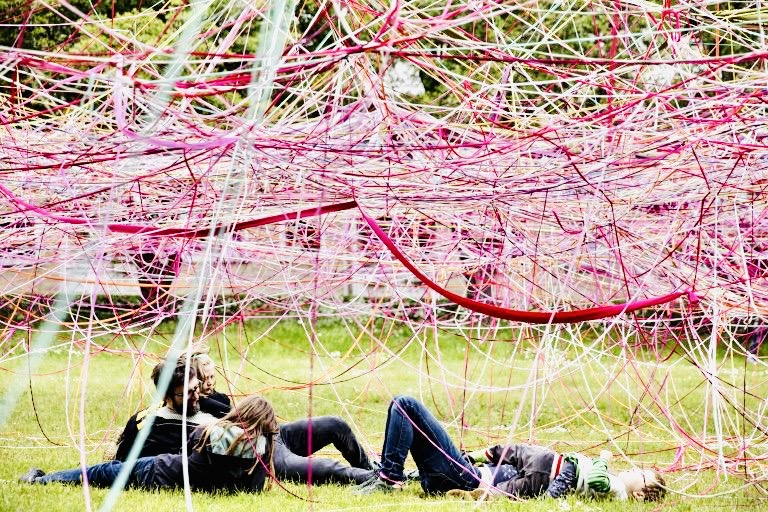 People lying on the grass beneath a low canopy woven from pink, red and white ribbons
